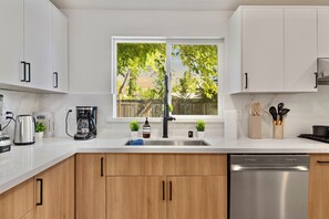 A modern kitchen with white cabinets, a stainless steel dishwasher, and a window with a view of trees. - 4BD 3BA Modern Home with Backyard. Near SF, Concord, Oakland & Napa Winery (Concord)