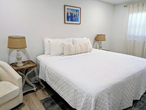 Een netjes opgemaakt bed met witte lakens, twee nachtkastjes met lampen, een ingelijst kunstwerk aan de muur en een raam met gordijnen.