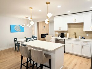 A modern kitchen with white cabinetry, a central island with bar stools, a dining area with blue chairs, and pendant lighting. - A Haven of Sun and Fun in Largo; A Private Oasis in the heart of everything (Largo)