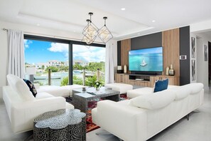 A modern living room with a large TV, white sofas, a coffee table, and a view of a body of water. - Rooftop Villa w/ Pool & Hot Tub in Chalk Sound (Providenciales)