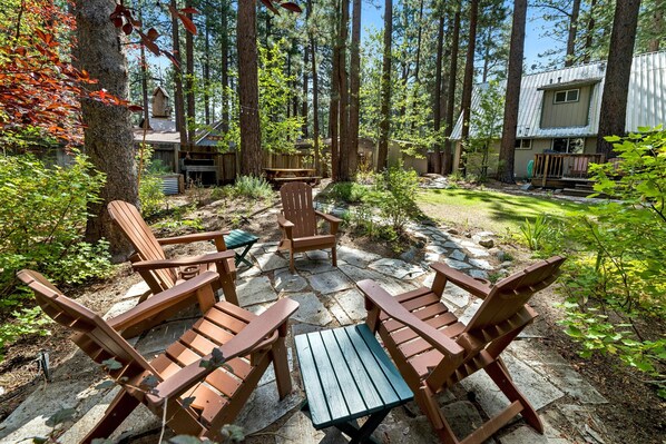 A patio area with Adirondack chairs and a wooden table, surrounded by a forested yard with a house in the background. - BTLR- Tahoe Pines Retreat | 4BR 2BA Cabin (South Lake Tahoe)