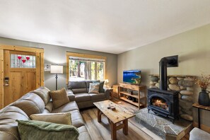 A cozy living room with a stone fireplace, a sofa, a coffee table, and a television. - BTLR- Tahoe Pines Retreat | 4BR 2BA Cabin (South Lake Tahoe)
