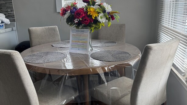Una mesa de comedor redonda con una cubierta de vidrio transparente, rodeada por cuatro sillas tapizadas. Un centro de mesa floral se encuentra en el centro de la mesa.