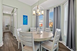 A dining area with a round table, white chairs, a chandelier, and grey curtains. - Seas The Magic | 2 Golf Carts | 4 Bikes | Walk to Pool | Near Dune Allen Beach (Santa Rosa Beach)