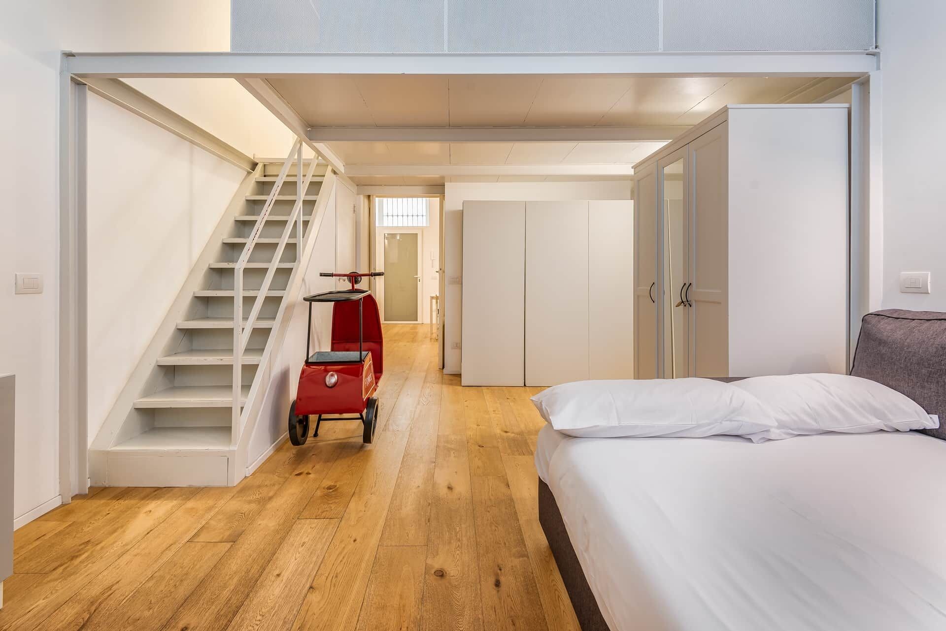 Un dormitorio con una cama, una maleta roja, una escalera y un armario.