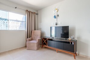 Ein modernes Wohnzimmer mit Fernseher, gepolstertem Sessel, schwarzem Unterhaltungsmöbel und einem Fenster mit Blick auf Gebäude.