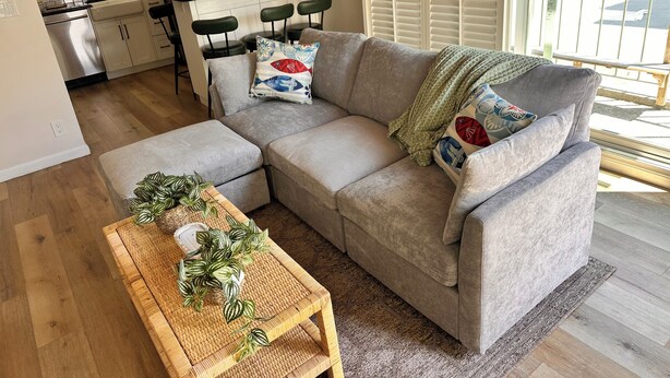 Een woonkamer met een grijze bank, een houten salontafel met een plant, en een keuken met een koelkast en fornuis.