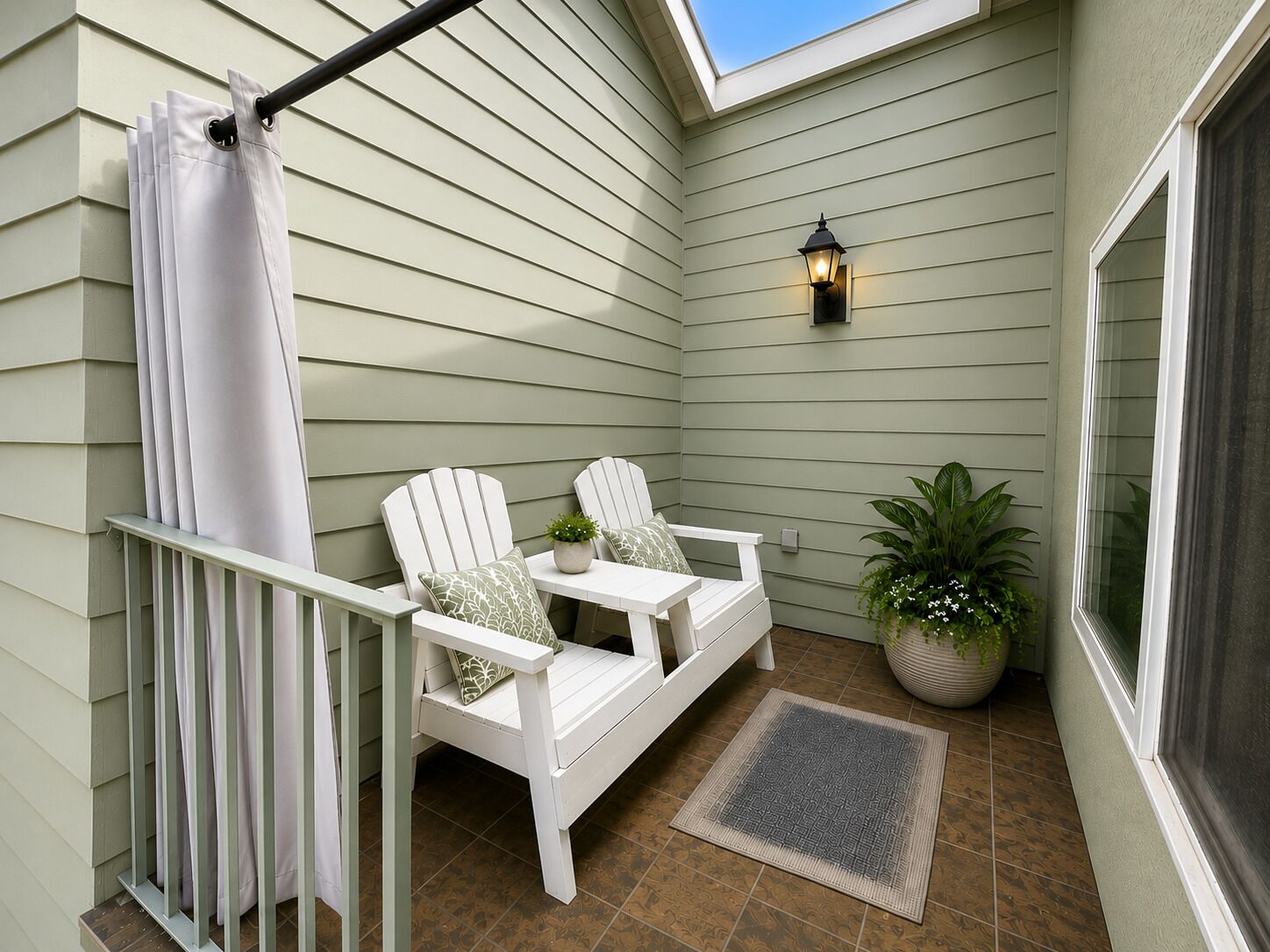 A small patio area with two Adirondack chairs, a white table, a potted plant, and a wall-mounted light fixture.