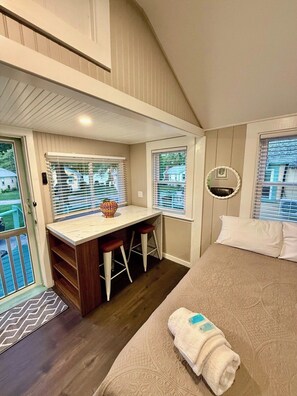 A compact bedroom with a bed, a small desk with stools, a window, and a door leading to an outdoor area. - Cabin 23 at Historic Sandy Point w. Boat Slip (Alton)