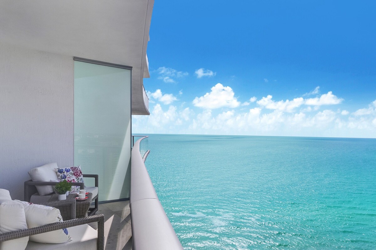 A balcony with a view of the ocean, a wicker chair, a small table with a plant, and a white cushion.