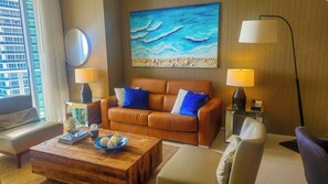 A living room with a brown leather sofa, two blue pillows, a wooden coffee table, and a painting of the ocean on the wall. - Lyfe Resort & Residences (Hollywood)