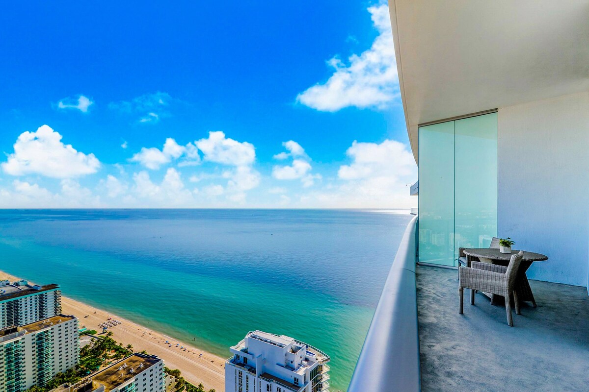 A balcony with a view of the ocean and beach, a small table with chairs, and a potted plant.