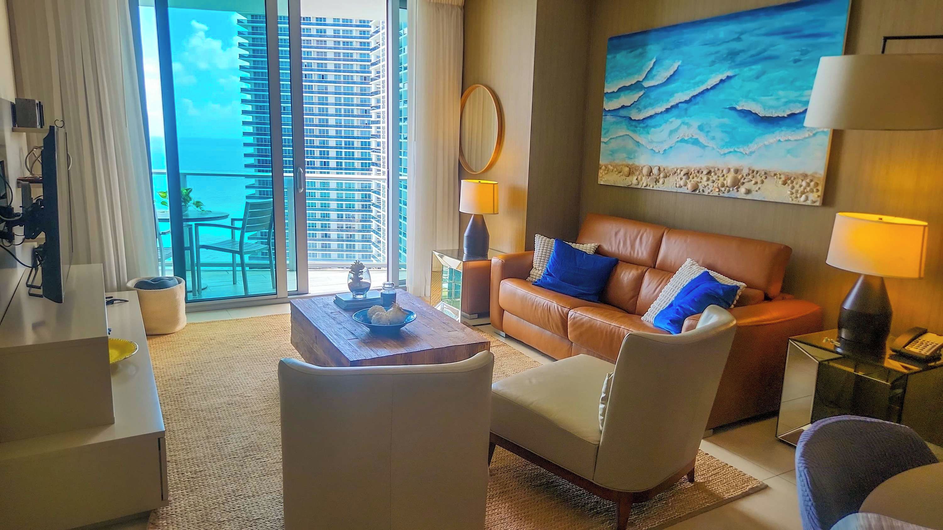 A hotel room with a leather sofa, a wooden coffee table, a round mirror, a painting of the ocean, and a view of a high-rise building.