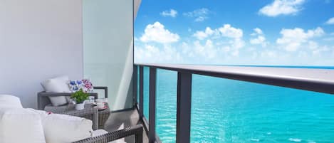 A balcony with a view of the ocean, white cushions, a wicker chair, and a small table with a plant.