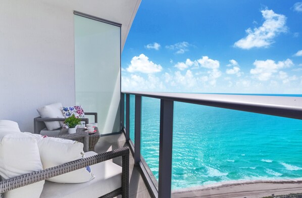 A balcony with a view of the ocean, white cushions, a wicker chair, and a small table with a plant.