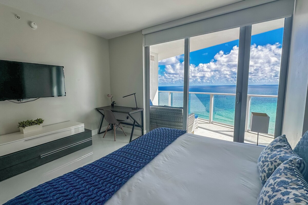 A modern hotel room with a large bed, a flat-screen TV, a desk with a chair, and a balcony with a view of the sea.