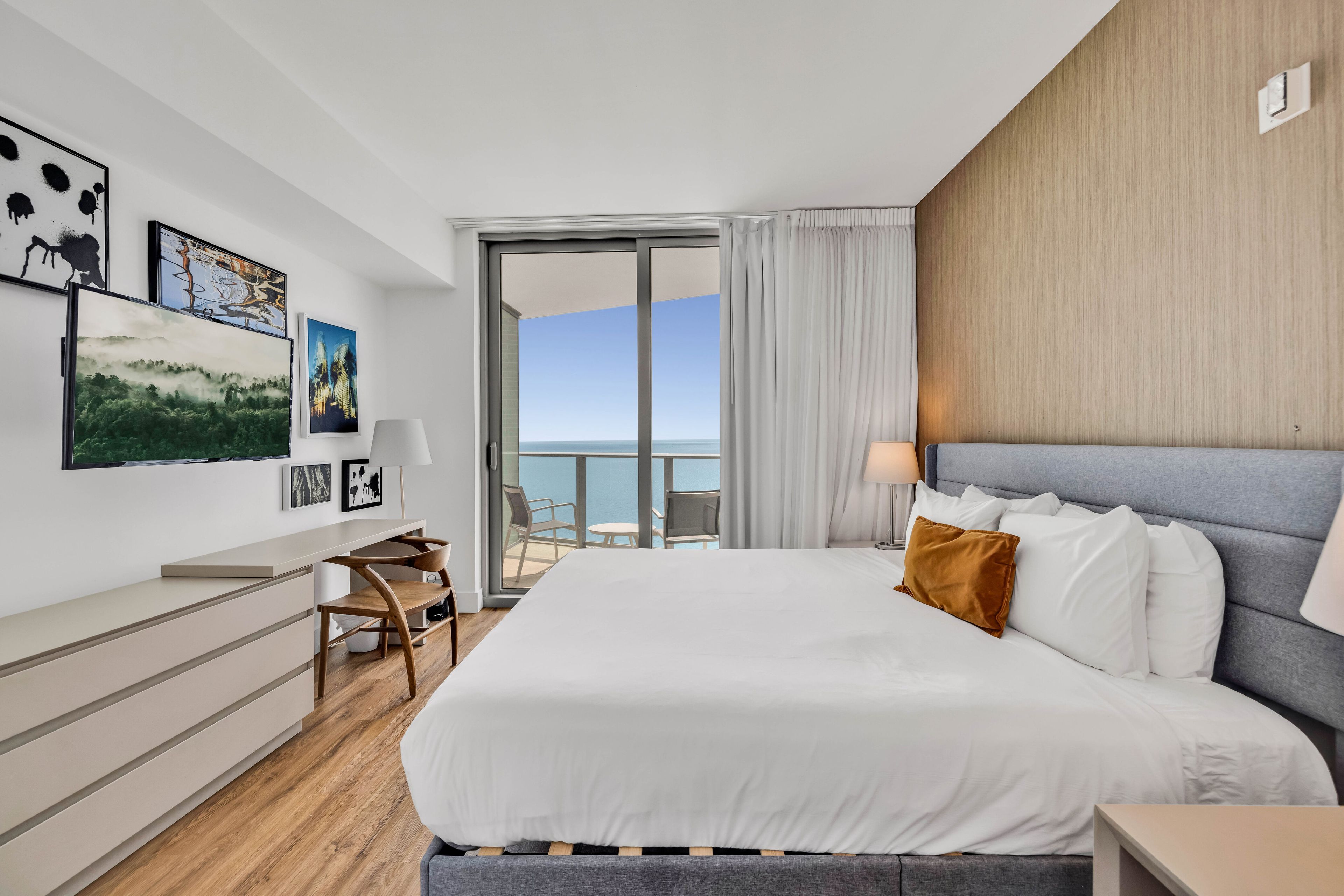A modern hotel room with a large bed, a desk, and a balcony with a view of the sea.