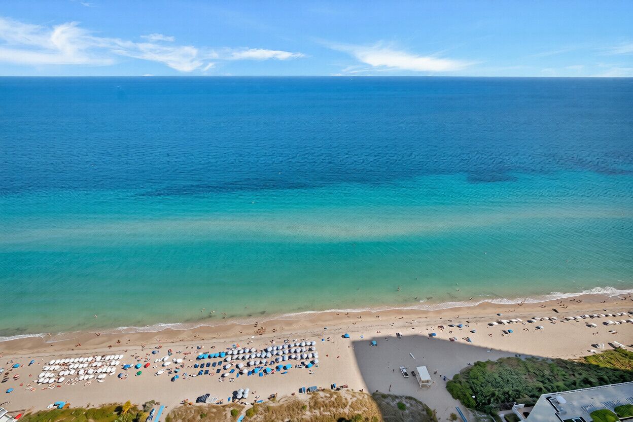 An aerial view of a beach with a clear sky, calm sea, and a sandy shore with umbrellas and lounge chairs.