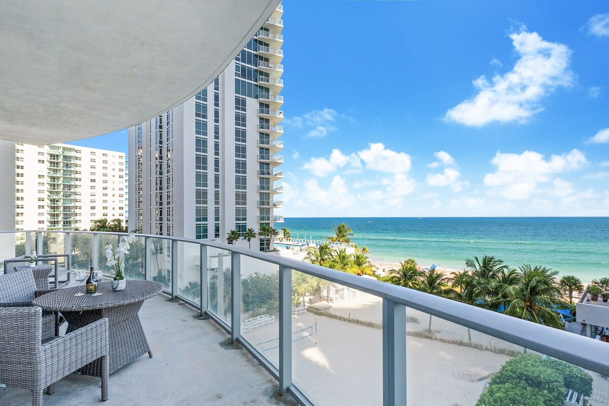 A balcony with wicker furniture, overlooking a beach and ocean with high-rise buildings in the background.
