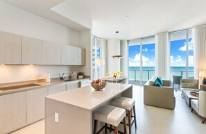 A modern kitchen with a central island, a dining area with a view of the sea, and a living room with a sofa and chairs.