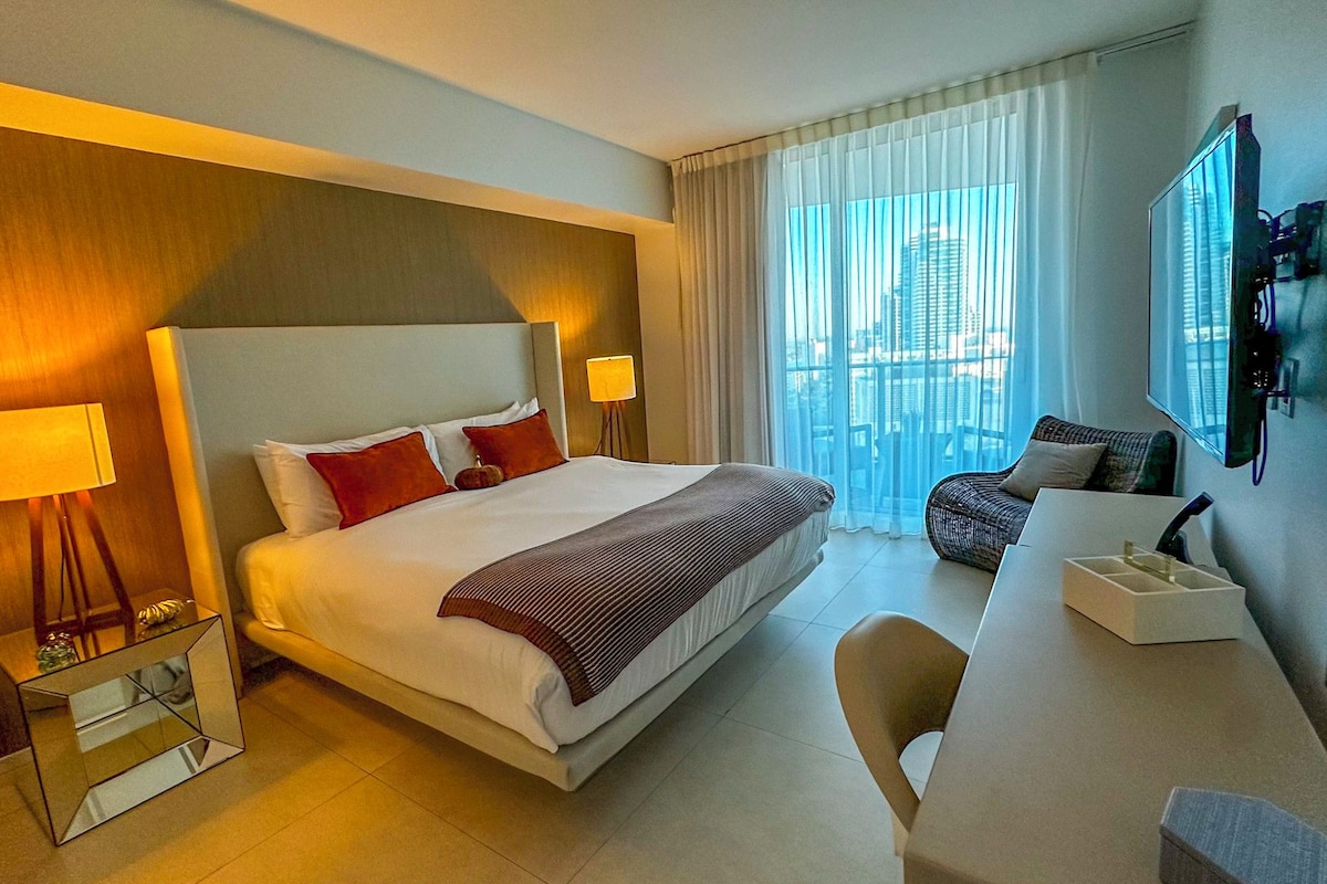 A hotel room with a large bed, bedside lamps, a sofa, a TV, and a city view.