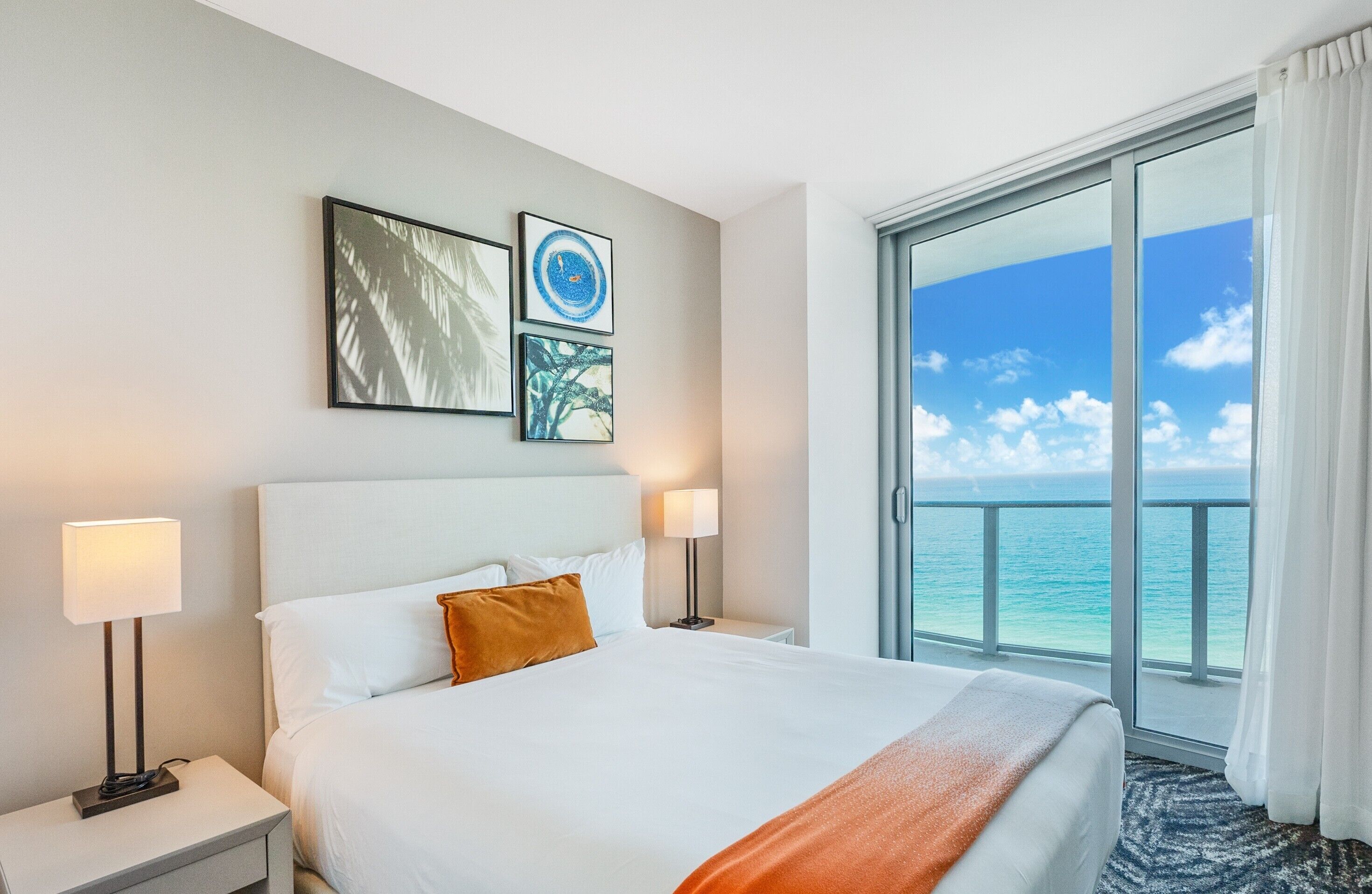 A hotel room with a bed, two bedside lamps, a nightstand, and a large window with a view of the ocean.