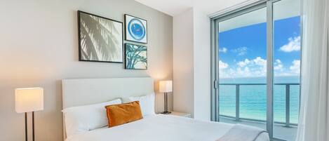 A hotel room with a bed, two bedside lamps, a nightstand, and a large window with a view of the ocean.