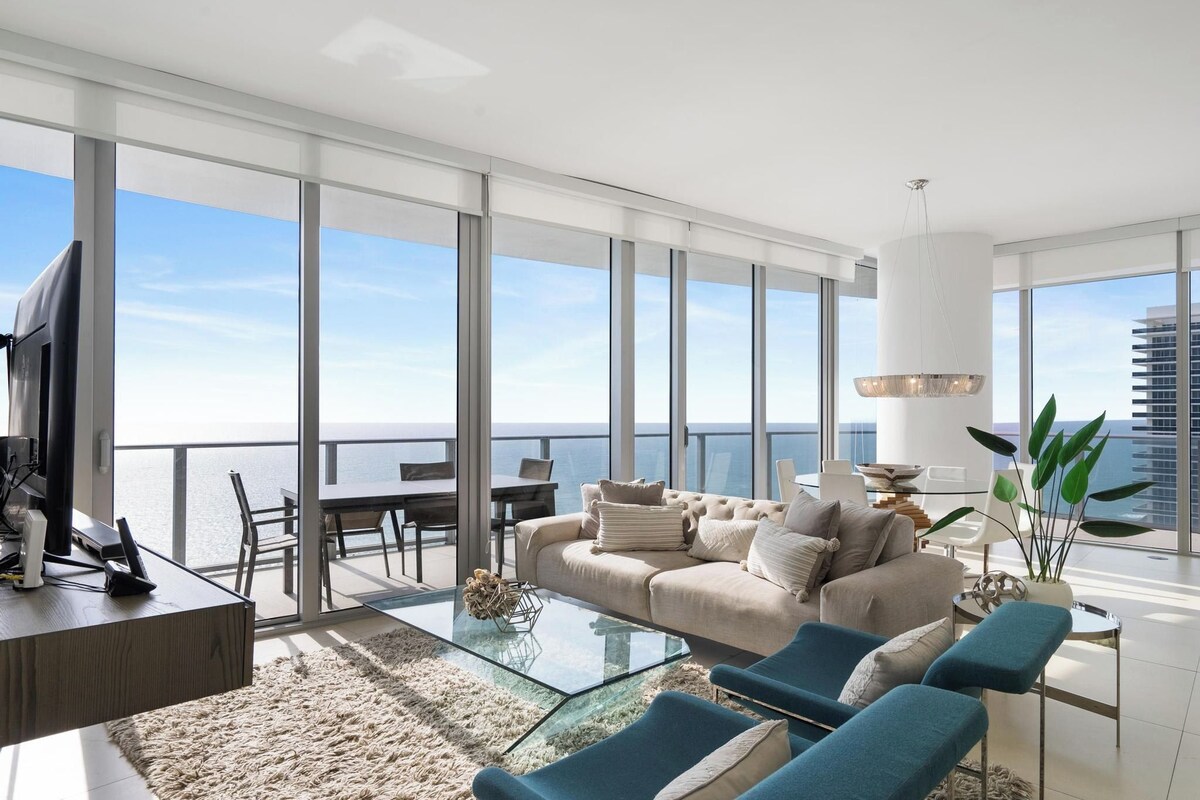 A spacious living room with a large sofa, a coffee table, and a TV. There is a balcony with a view of the ocean.