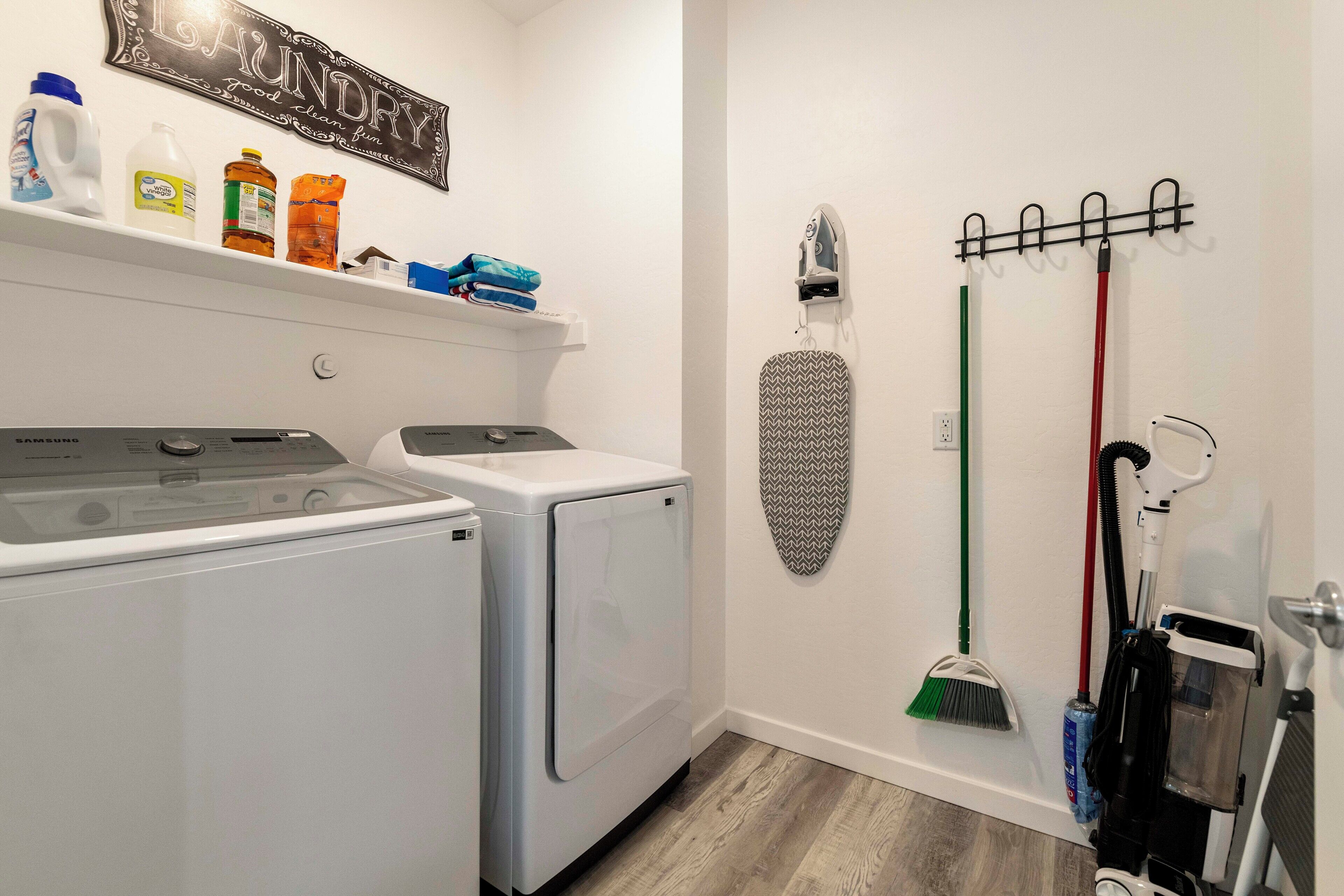 A laundry room with two front-loading washers, a drying rack, a wall-mounted ironing board, and cleaning supplies.