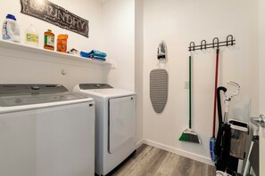 Una sala de bugaderia amb dues rentadores de càrrega frontal, un estenedora de roba, una taula de planxar penjada a la paret i productes de neteja.