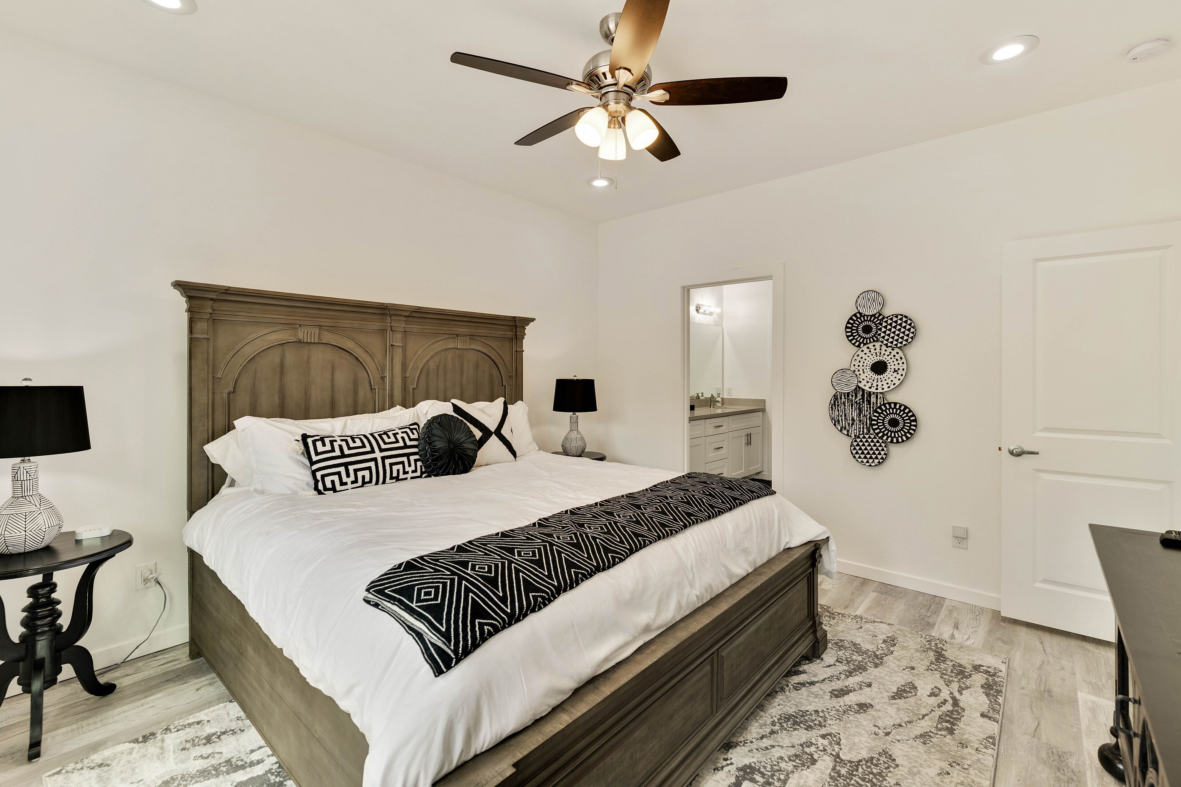 A bedroom with a bed, a ceiling fan, a nightstand, a lamp, a mirror, and wall art.