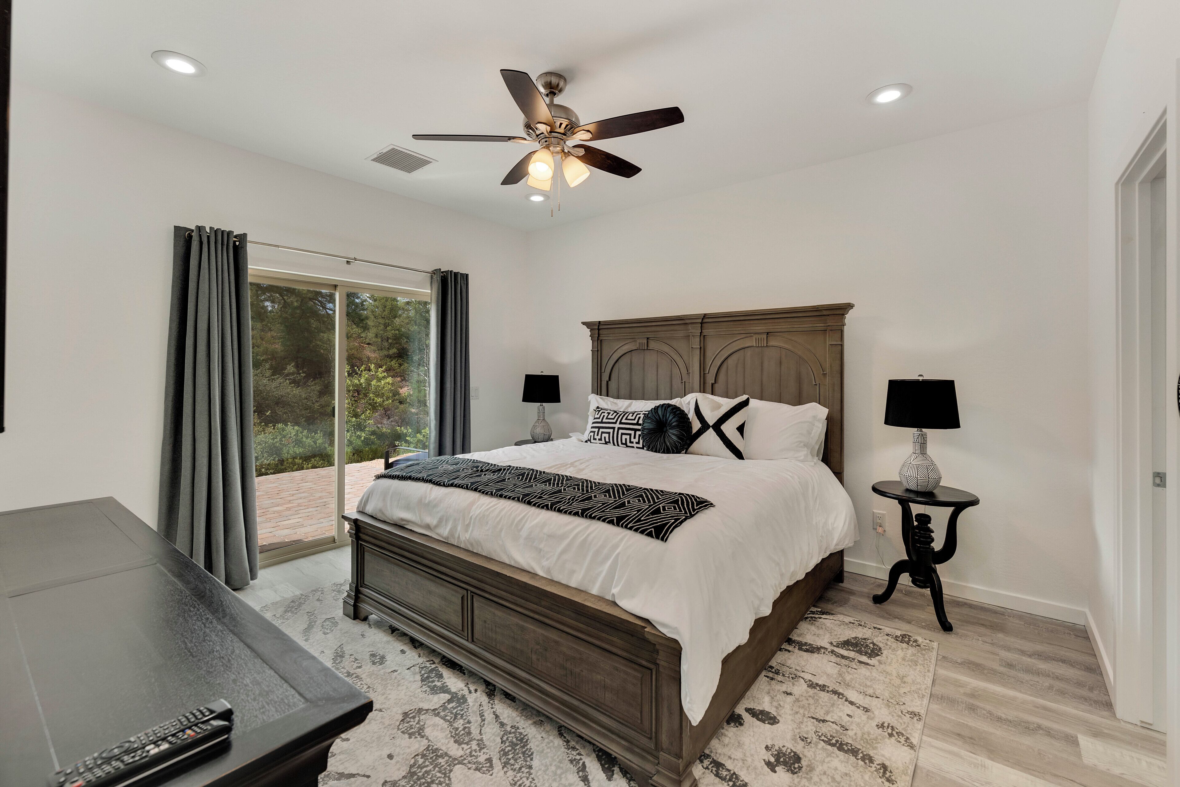 A bedroom with a large bed, a ceiling fan, a nightstand, and a view through a window.
