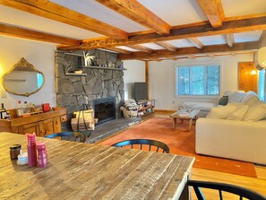 A cozy living room with a stone fireplace, wooden beams, a sofa, and a dining area with a table and chairs.