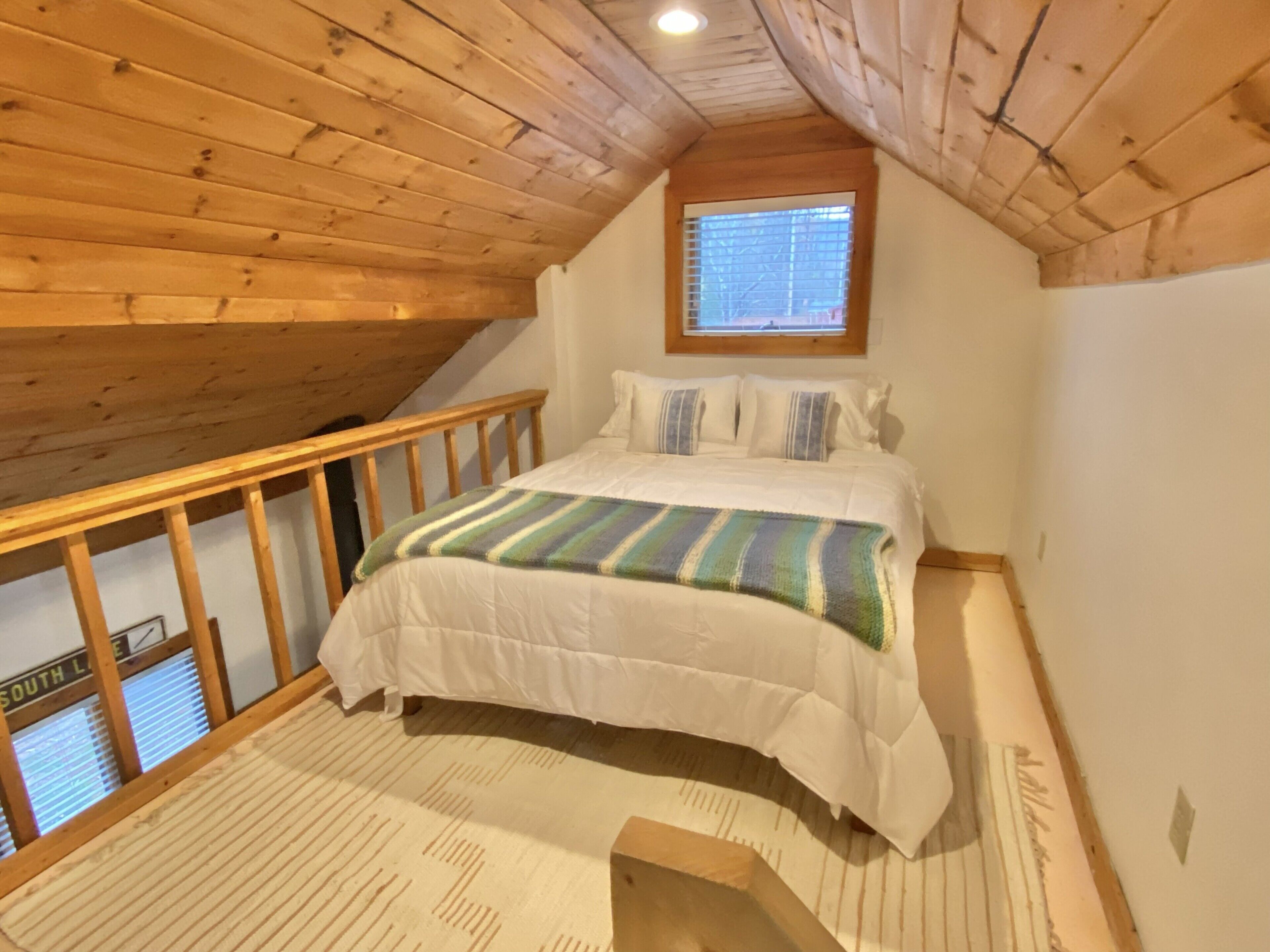 A bedroom with a wooden ceiling, a window, a bed with a striped blanket, and a view of the outside.