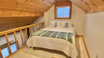 A bedroom with a wooden ceiling, a window, a bed with a striped blanket, and a view of the outside.