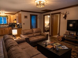 A living room with a sofa, a coffee table, and a television. There is a kitchen area with cabinets and a microwave.