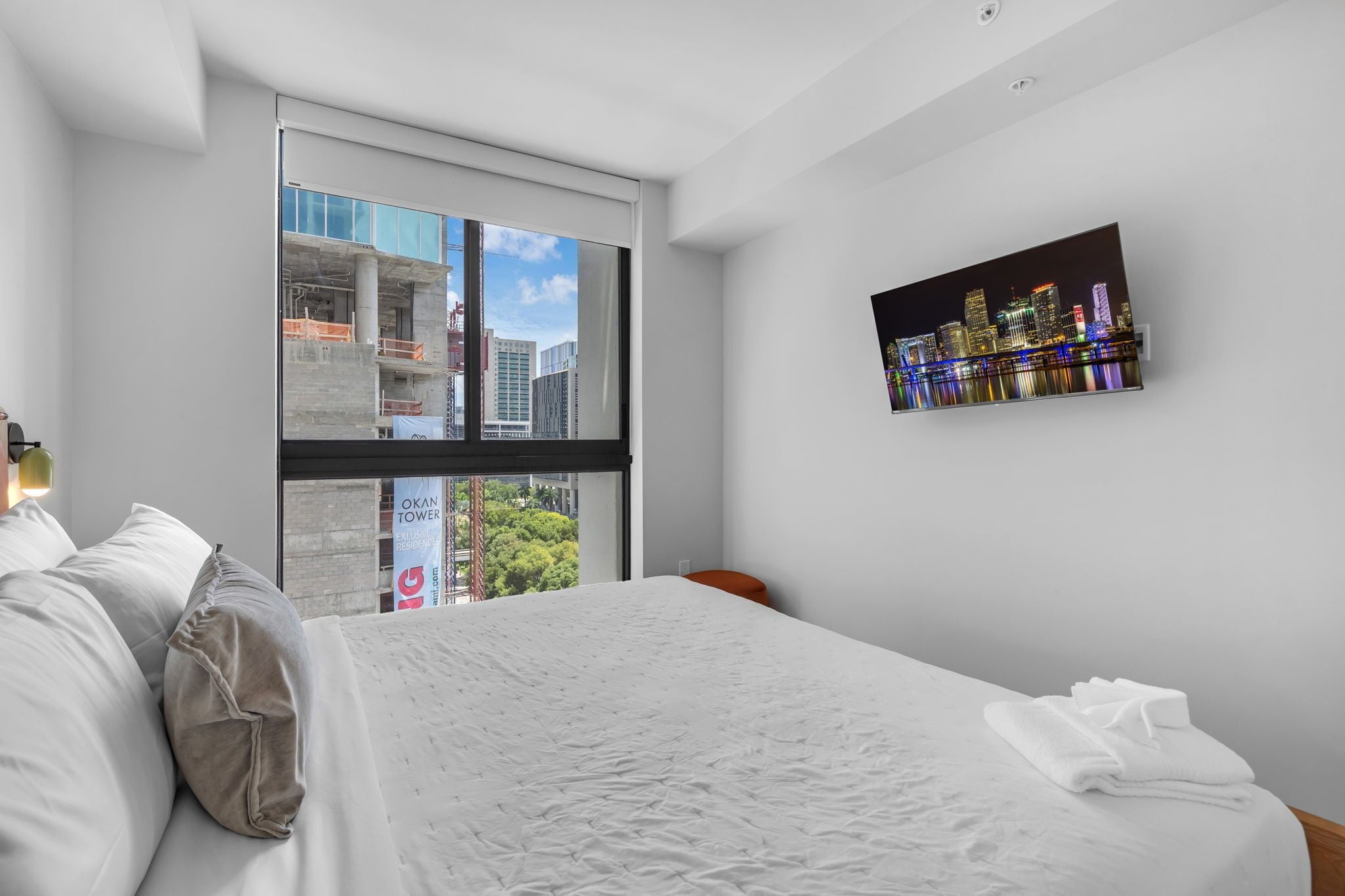 A modern hotel room with a large bed, a cityscape view through a floor-to-ceiling window, and a framed cityscape artwork on the wall.