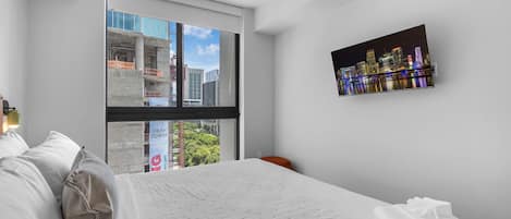 A modern hotel room with a large bed, a cityscape view through a floor-to-ceiling window, and a framed cityscape artwork on the wall.