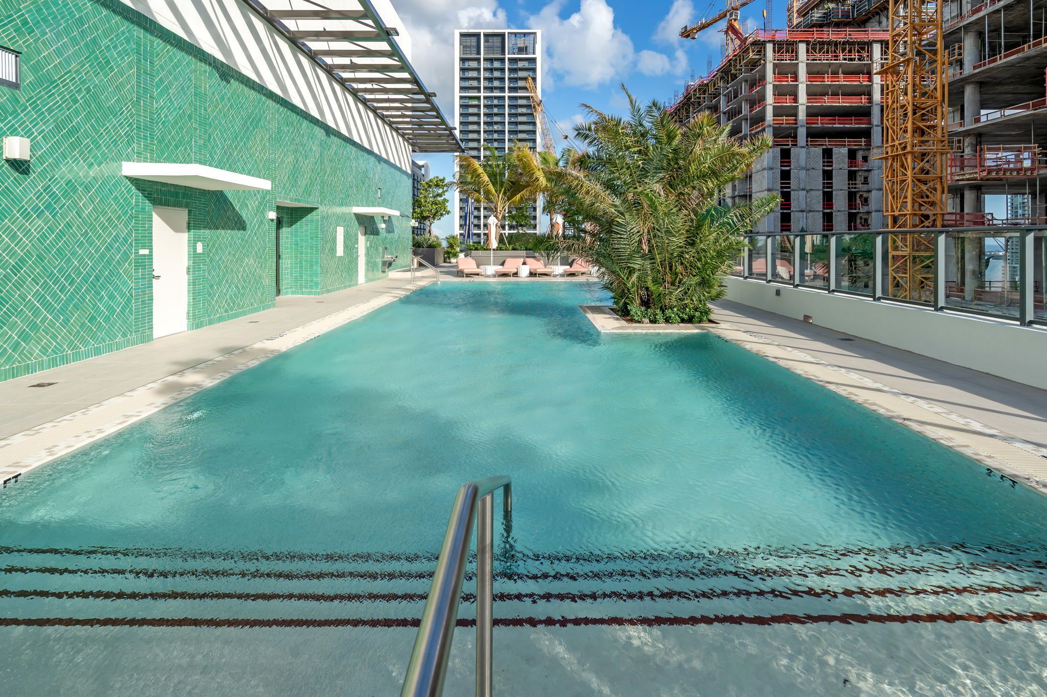 Une piscine à l’eau claire, entourée d’un bâtiment dont la façade est recouverte de carreaux verts, avec un chantier de construction en arrière-plan.