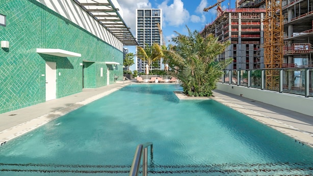 A swimming pool with clear water, surrounded by a building with a green tiled exterior and a construction site in the background. - The Crosby Miami Worldcenter (Miami)