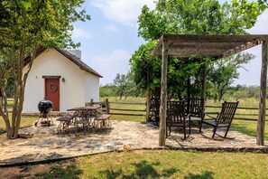 Un cadre extérieur rustique avec une terrasse en pierre, une pergola en bois et un petit bâtiment blanc doté d’une porte rouge.