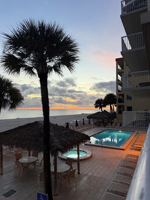 A pool area with lounge chairs and a thatched umbrella, overlooking a beach and ocean. - Oceanfront Paradise in Madeira Beach. Private Balcony.  Pool & Hot Tub (Madeira Beach)