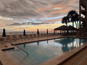 A swimming pool with lounge chairs and umbrellas, a fence, and a view of the sunset over the ocean. - Oceanfront Paradise in Madeira Beach. Private Balcony. Pool & Hot Tub (Madeira Beach)