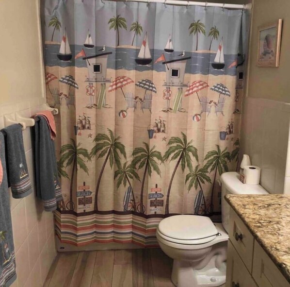 A bathroom with a beach-themed shower curtain, a toilet, and striped towels. - Oceanfront Paradise in Madeira Beach. Private Balcony. Pool & Hot Tub (Madeira Beach)
