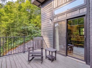 Eine Holzterrasse mit Schaukelstuhl und Tisch, mit Blick auf ein bewaldetes Gebiet.