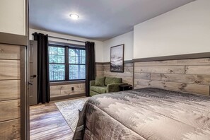 Een slaapkamer met een groot bed, een groene fauteuil, een klein tafeltje en een raam met gordijnen.