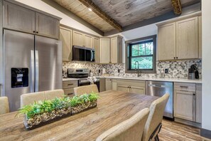 A modern kitchen with stainless steel appliances, a stone backsplash, and a large dining table with chairs. - Bear Trap in the Glades (Gatlinburg)