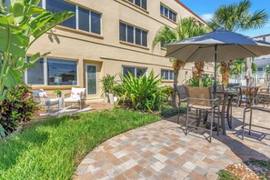 A patio area with outdoor furniture, a large umbrella, and a building with multiple windows. - NEW! Flamingo Breeze Waterfront • Walk to Beach and Restaurants (Treasure Island)