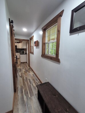 A narrow hallway with wooden flooring, a bench, and a window with a view of greenery.