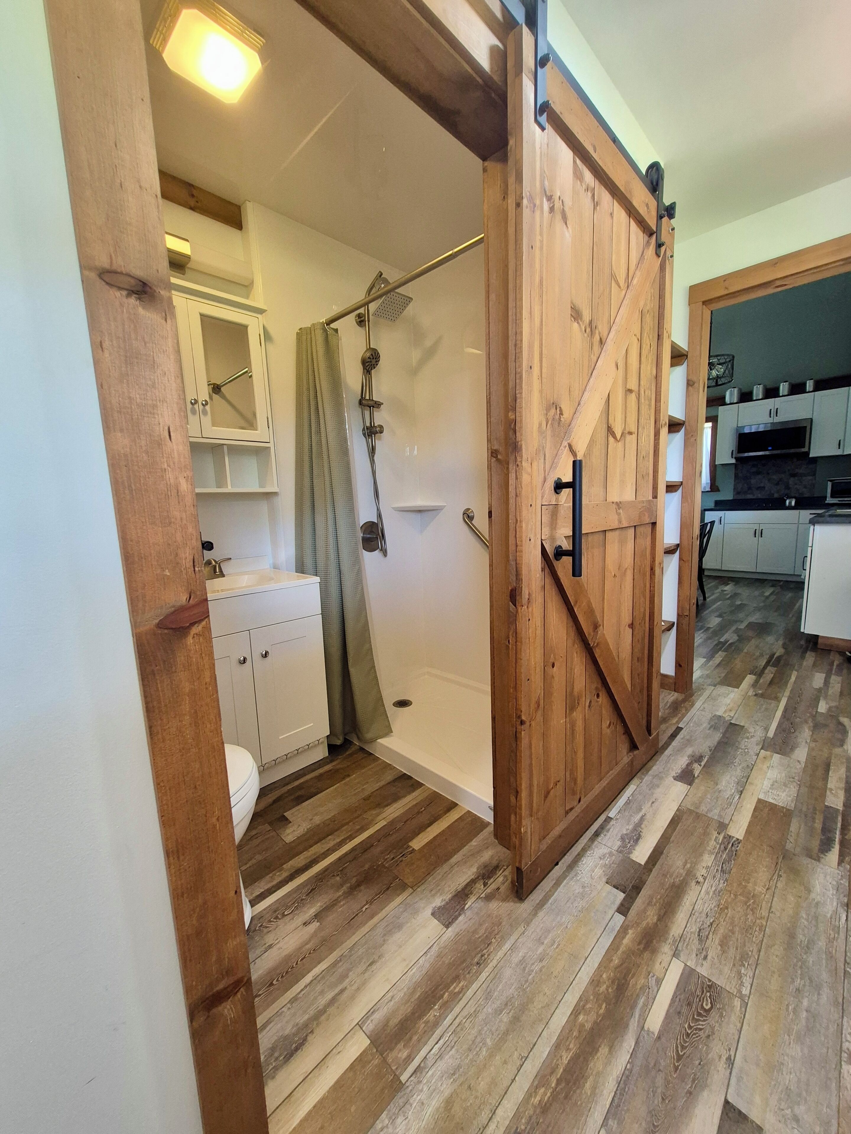 A bathroom with a shower, toilet, and a wooden door leading to a kitchen area.
